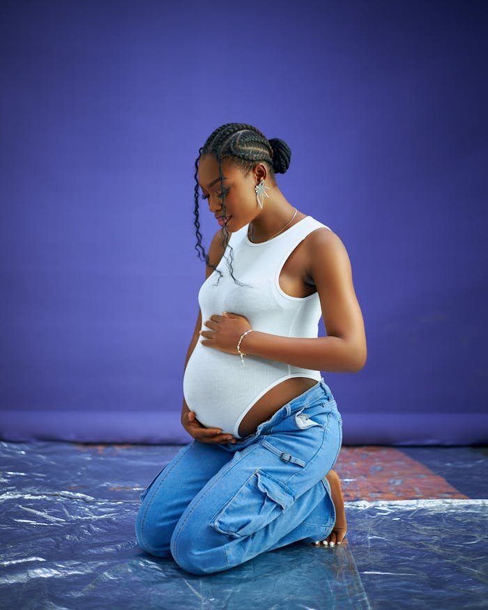 Portrait of a pregnant woman kneeling, wearing a white top and blue jeans against a purple backdrop.